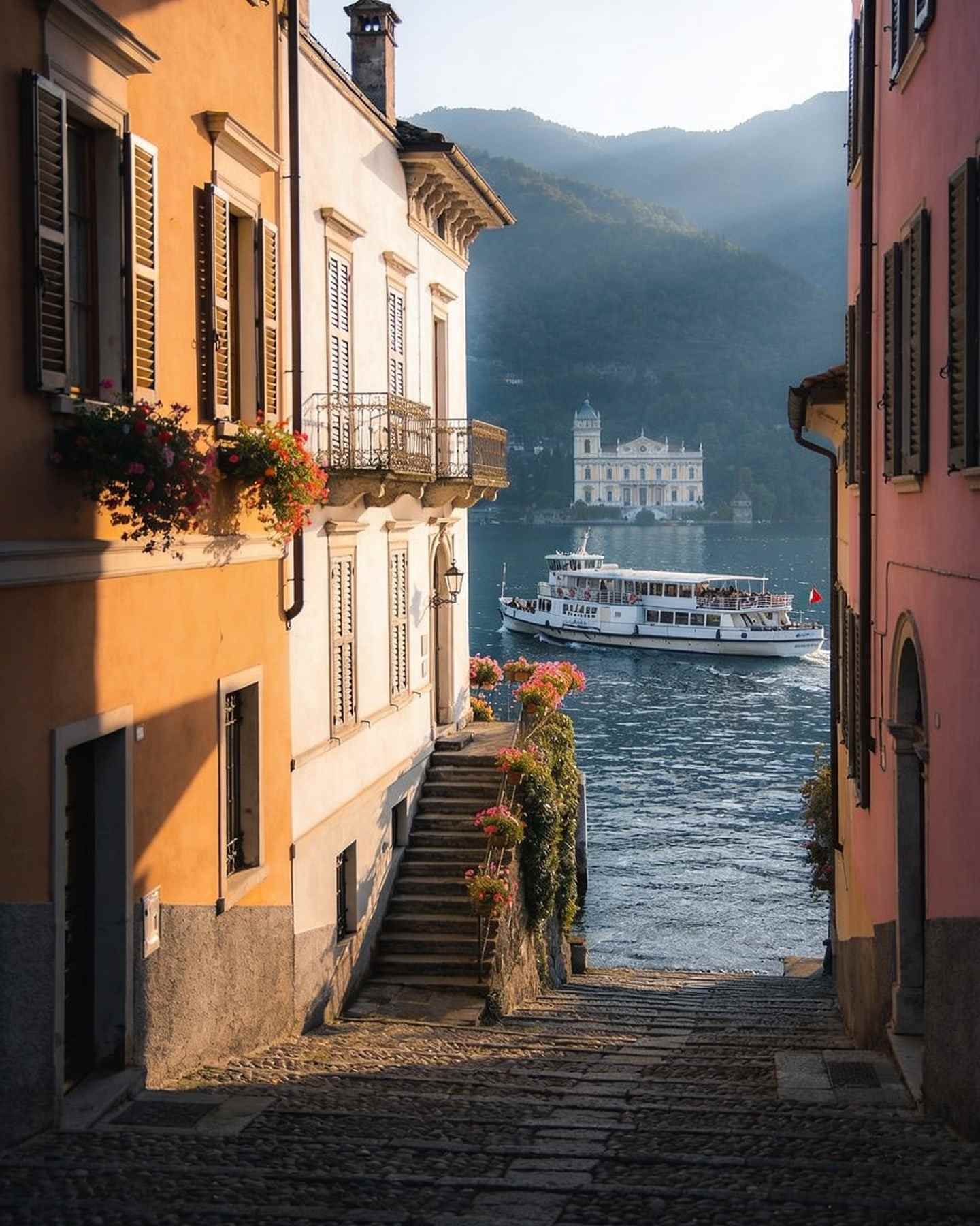 Ochre and pink houses along a lakeside promenade, cobbled steps climbing between them and a ferry arriving.