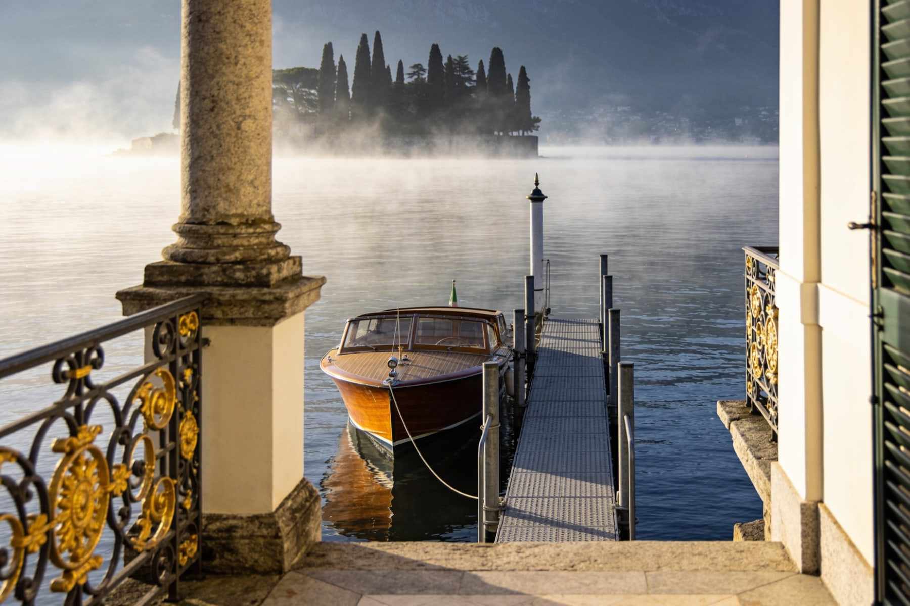 Morning mist on the lake seen from a villa jetty with a wooden boat moored and cypress silhouettes.